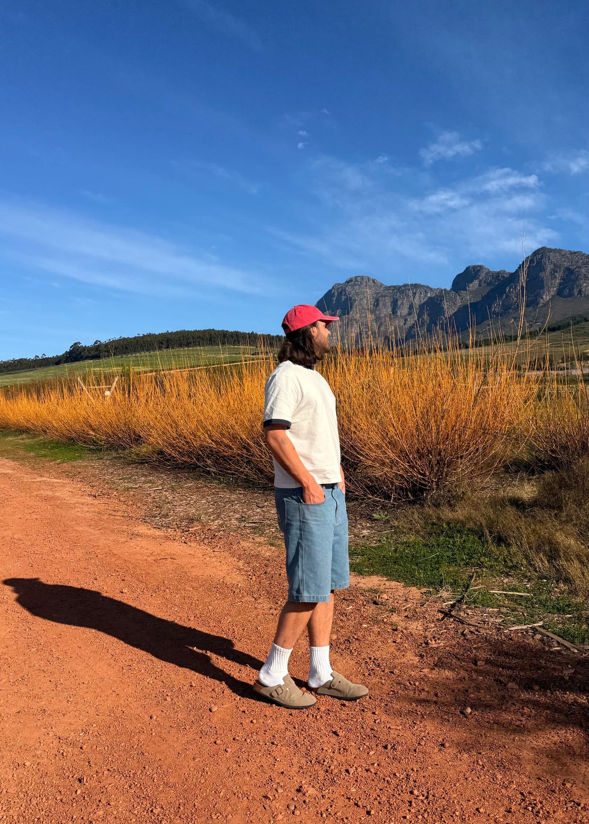 A man wearing a pink cap, white t-shirt, and denim shorts stands on a dirt path in a rural landscape with dry grass and mountains under a blue sky.